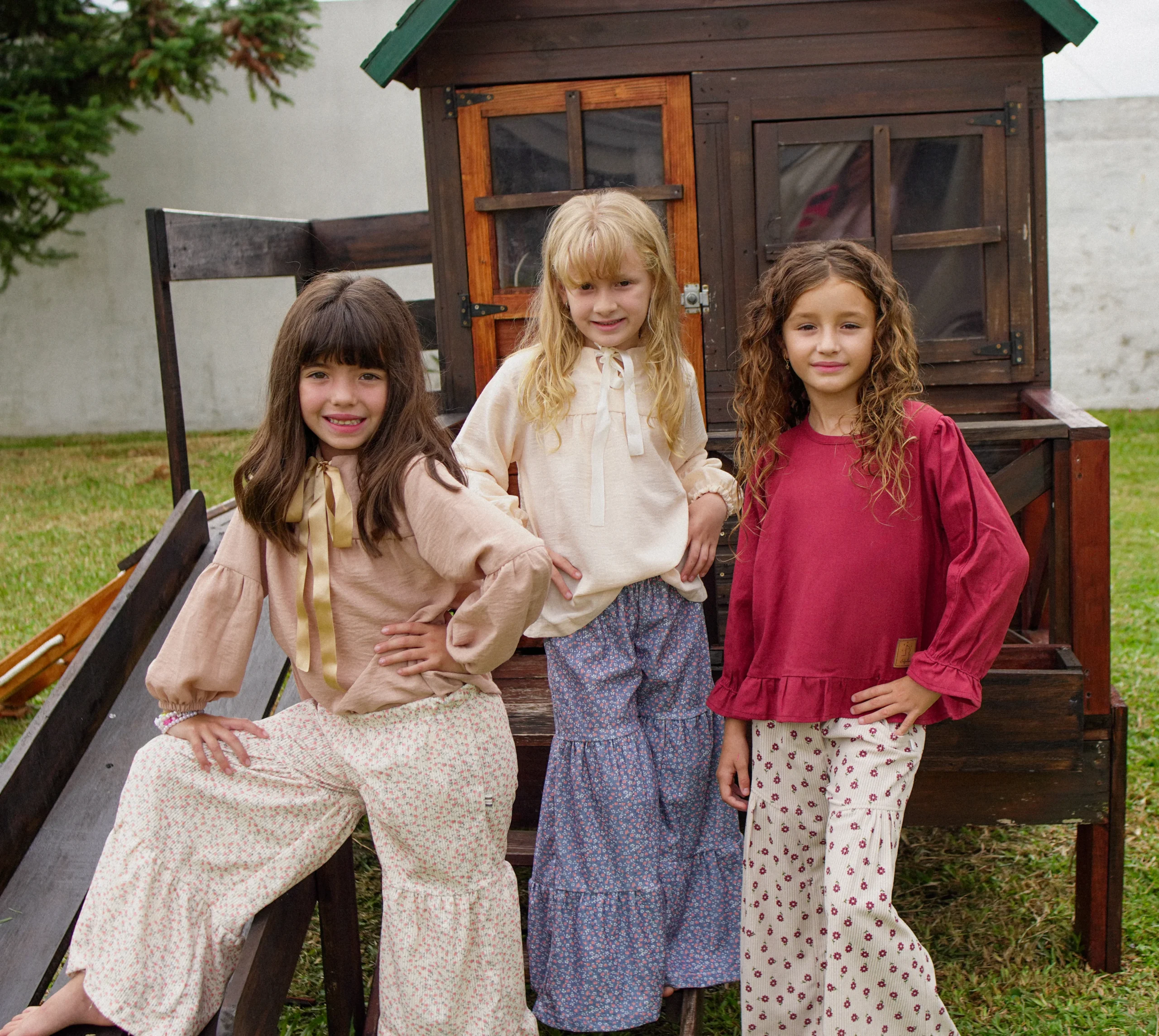 Tres niñas sentadas en casita de madera con blusas crema y faldas estampadas Infantem